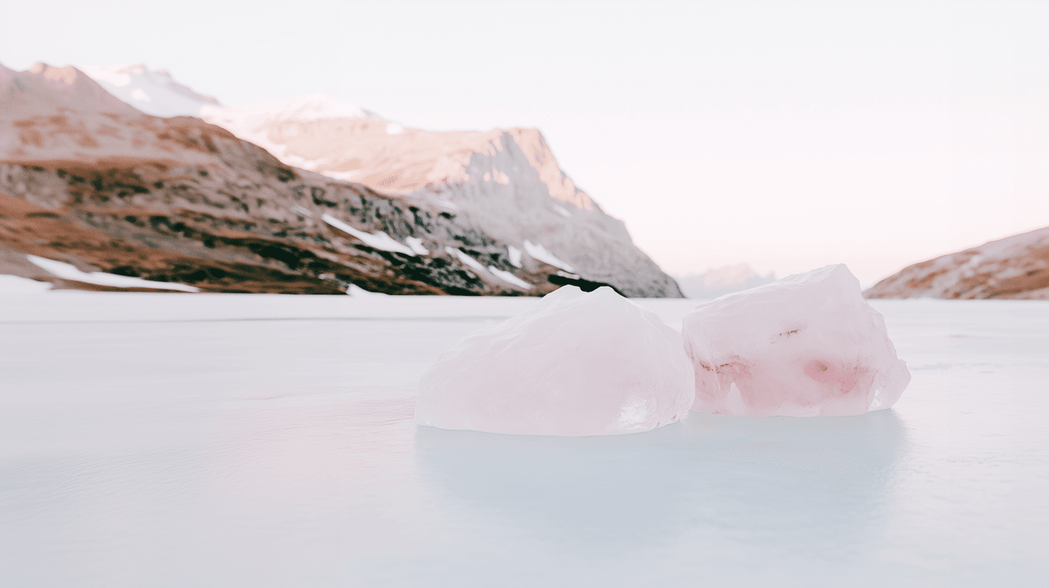 Pink-tinged ice on a frozen lake with snow-capped mountains behind